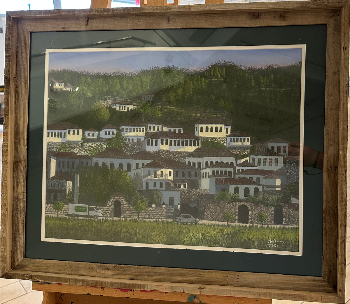 Framed artwork of a scenic village on an easel in a gallery setting